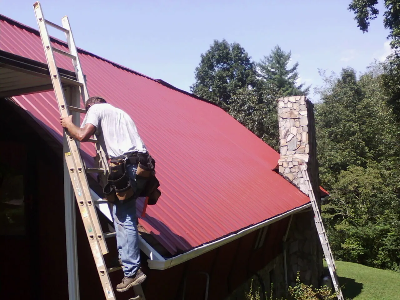 Roofer on a ladder beside a red metal roof and stone chimney — Soffit & Fascia Repair by Roofing Brothers in Trimble